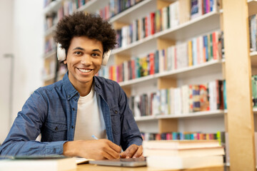 Portrait of happy black student guy wearing headphones and taking notes in front of laptop in...