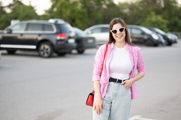 young woman wearing pink and white plaid shirt print vichy smiles while standing parking lot. white cat eyes sunglasses and red handbag casual outfit. fashionable stylish look of the day 