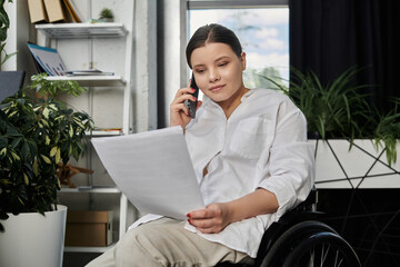 A young businesswoman in a wheelchair works on documents and talks on the phone in a modern office.