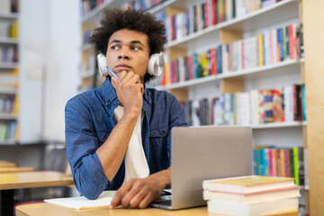 Pensive African American male student in headphones looking away, creating essay or working on online project sitting with laptop and books in university library
