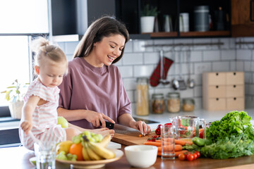A cheerful mother is making a healthy meal with lots of fruit and vegetables, and her cute little baby daughter is keeping her company