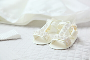 White baby shoes decorated with pearls lying on a christening gown on a white quilted blanket