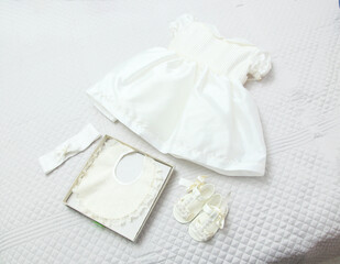 White ceremony dress, shoes, bib, and headband laid out on a bed for a baby girl, capturing...
