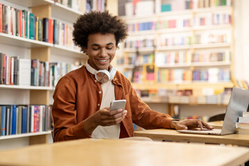 African American male student using cellphone and laptop sitting at desk in college library interior, texting while preparing for exams