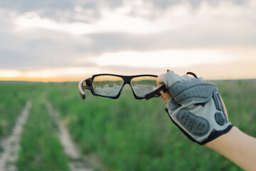 A woman wearing a cycling helmet holds up a pair of cycling glasses in front of her face. She is wearing a blue shirt and black cycling gloves.