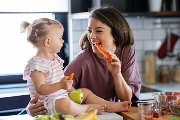 Cheerful mother and her cute little baby are eating healthy fruit and vegetables in the home kitchen