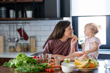 Cheerful mother and her cute little baby are eating healthy fruit and vegetables in the home kitchen