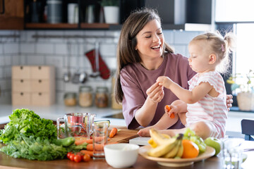 Cheerful mother and her cute little baby are eating healthy fruit and vegetables in the home kitchen