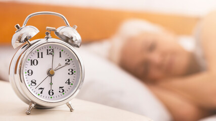 An alarm clock sits undisturbed on top of a neat bed, display showing the time over sleeping senior...