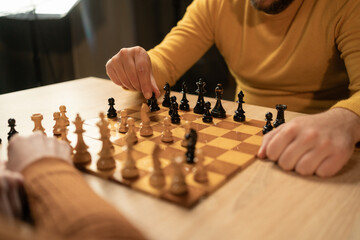 Close-up view of two men playing chess at home. The focus is on their hands and the wooden...