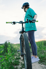 A woman wearing white sneakers stands next to a mountain bike with a black tire on a dirt path. The bike is parked, and the woman is looking off-camera.