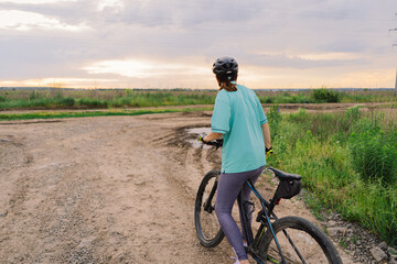 Fototapeta premium A woman wearing a helmet rides a black mountain bike along a dirt path. The path runs through an open grassy field, with houses and trees visible in the background.
