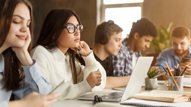 A collection of multiethnic students is deeply focused on their individual studies at a long table basked in sunlight. With books open and laptops out, they sit intently working, some writing notes