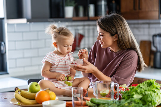 The mother prepared a healthy fruit and yogurt meal rich in vitamins, and she's feeding her cute little baby daughter at the home kitchen counter