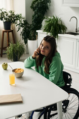 A young brunette woman with a disability sits in a wheelchair at her kitchen table, talking on her cell phone.