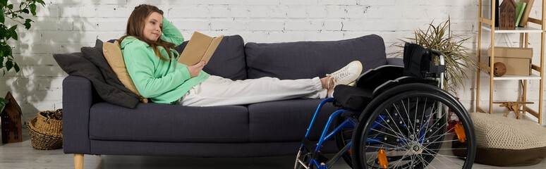 A brunette young woman sits in a wheelchair, resting her legs on a couch while reading a book in...