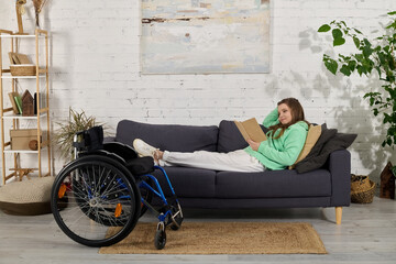 Obraz premium A young brunette woman with a disability relaxes on a sofa in her living room, reading a book. Her wheelchair sits beside her.