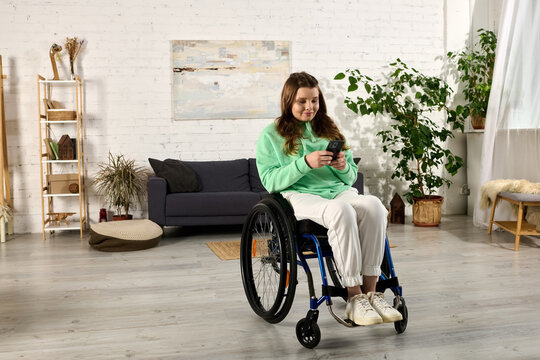 A young brunette woman in a wheelchair is using her phone while sitting in her living room.