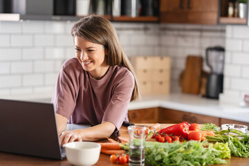 A young, pretty, cheerful woman is looking at a laptop computer while preparing food at home, following a cooking recipe online