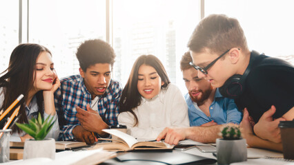 A diverse group of students are gathered around a wooden table, engrossed in a book placed in the center. Some are pointing at the pages, while others are consulting additional materials