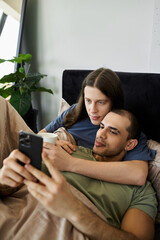 A young gay couple relaxes in bed, embracing and looking at a phone together.