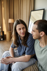 A young gay couple sits together in a bedroom, sharing a tender moment.