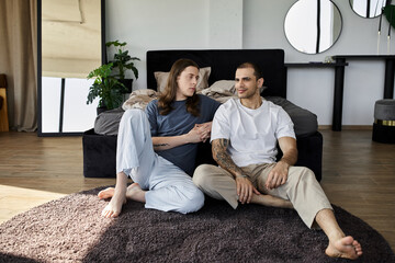 Two young men sit on a plush rug in a modern bedroom, engaged in conversation and enjoying each...