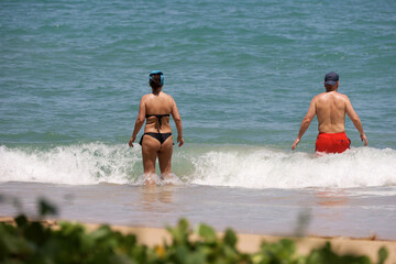 Couple going to swim in tropical sea. Woman in black bikini and guy in red shorts walk together by...