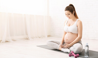 A pregnant woman is seated on a yoga mat, engrossed in her phone. She appears focused as she scrolls through the screen. Her bump is prominent, indicating her stage of pregnancy.