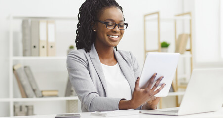 Smiling young African American businesswoman using a digital tablet, sitting at desk at modern office. Business app concept © Prostock-studio
