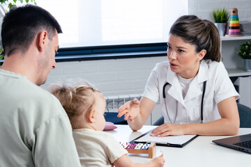 Fototapeta premium A woman pediatrician is examining her little girl patient and talking with her father