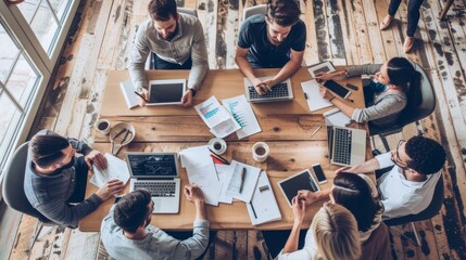 Top view of a creative team meeting around a wooden table, using laptops and tablets for a collaborative brainstorming session.