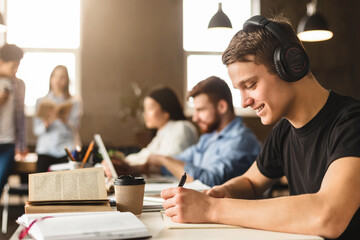 Student guy preparing for classes, taking notes in university library, listening music in headphones