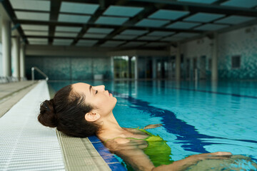 A young, beautiful brunette woman in a swimsuit floats effortlessly in an indoor spa swimming pool.