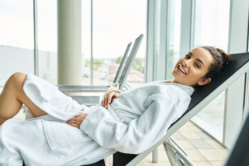 A young, beautiful brunette woman in a bathrobe sits peacefully in a lounge chair indoors at a spa...