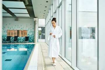 A young, beautiful brunette woman in a bathrobe standing beside an indoor swimming pool.