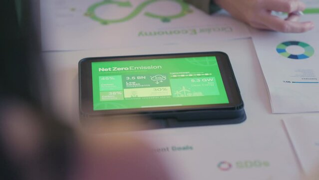 Close-up of business professionals analyzing net zero emission data on a tablet during a meeting focused on sustainability strategies.