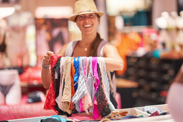 A joyful woman is seen smiling, holding colorful underwear at a market stall on a sunny day