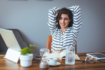 A smiling Caucasian woman in striped business attire stretches for relaxation amidst her busy office environment, surrounded by work essentials like a laptop and notepad.