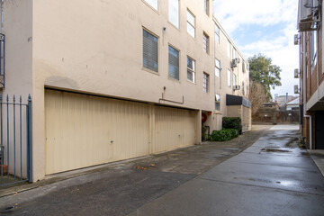 An urban alleyway between multi-story old residential apartment buildings with a row of large garage doors at the ground level in a suburb of Australia's neighborhood.