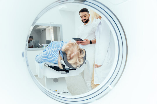 Radiographer operating computed tomography scanner being used to scan lying patient in radiology department of hospital. Doctor holding a tablet and looking at the patient. - Powered by Adobe