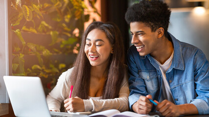 African American young guy and girl are seated at a table in a cafe, focused on a laptop screen. They appear to be engaged in collaborative work, possibly discussing or completing a project together.