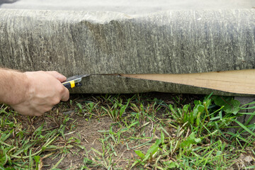 A hand carefully cuts through a piece of green and brown landscaping material with a utility knife....