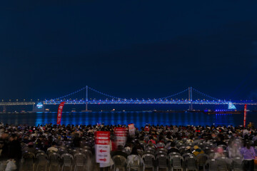 a night scene at Busan beach where a crowd of tourists eagerly awaits a fireworks display. The city&rsquo;s illuminated buildings create a stunning backdrop