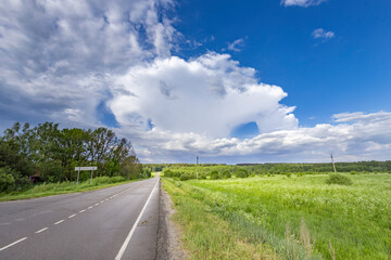 A picturesque scene of a country road winding through verdant fields under a vast, blue sky. Fluffy white clouds drift across the horizon, casting shadows on the green landscape.