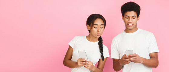 Cheating and infidelity. Girl spying and peeking at smartphone of her boyfriend, pink background