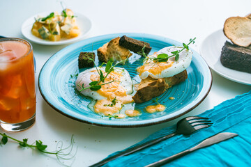 Poached eggs with hollandaise sauce on toast with crouton on a turquoise plate, next to a glass of orange juice and cutlery