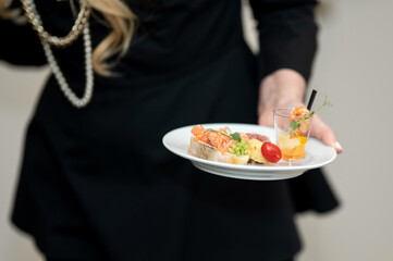 woman holds plate of snacks. Festive buffet menu. Corporate catering. Shrimp appetizers in glass. girl in black clothes holds plate of food. Front view. Light background. High quality photo