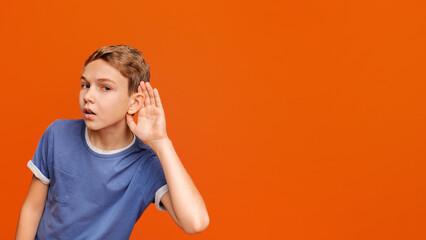 Curious snoopy schoolboy holding hand at his ear, trying to overhear private conversation, posing at orange studio background