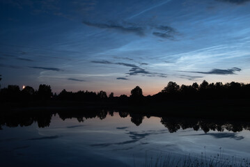 silver clouds reflected in the lake on a summer night with the moon above the trees